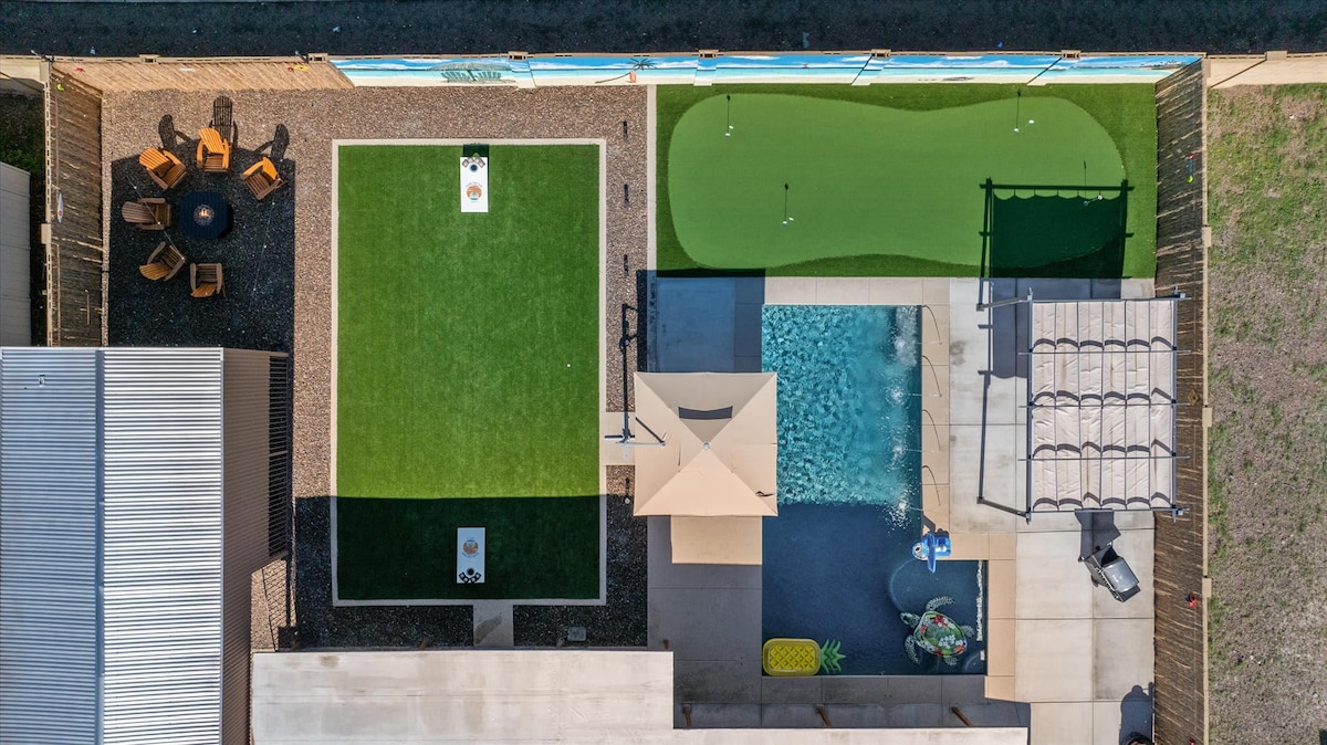 An aerial view of the outdoor area features a heated saltwater pool, a spacious putting green, and a lawn for outdoor games. A shaded patio with seating surrounds a fire pit, providing a cozy outdoor space for relaxation and entertainment.