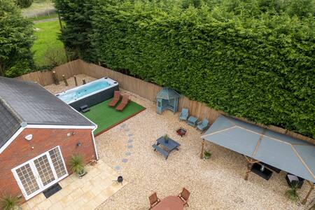 An aerial view captures a spacious outdoor area featuring a Swim Spa, surrounded by a manicured hedge. Comfortable seating arrangements are visible, including lounge chairs and a shaded gazebo. A gravel patio area includes a fire pit with additional seating, inviting relaxation and outdoor gatherings.