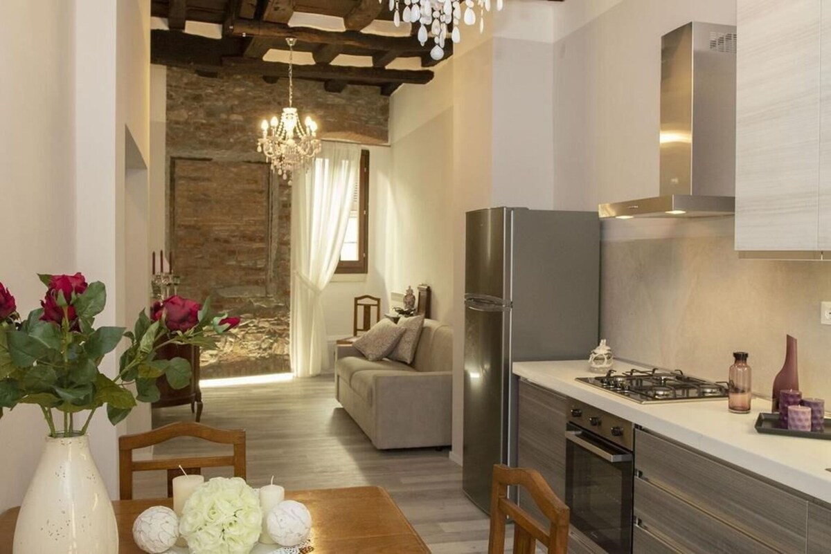 A cozy living area features a light-colored double sofa bed beside a fully equipped kitchenette. The space is enhanced by wooden beams on the ceiling and a chandelier. A dining table with floral arrangements is visible, along with a glimpse of a well-lit area in the background.