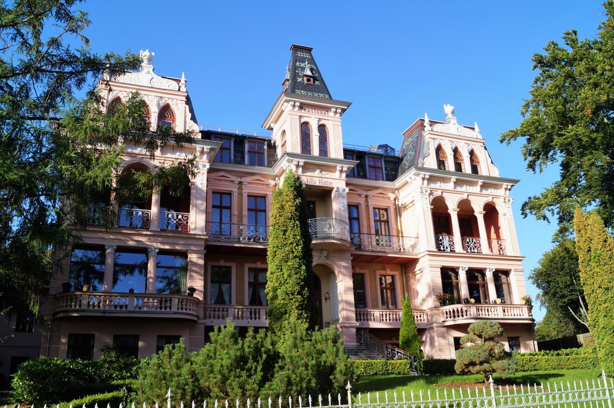 The villa features an elegantly ornate façade with intricate architectural details. Lush greenery surrounds the building, while large windows provide an inviting view. The structure is complemented by a taller central tower, conveying a sense of grandeur against a clear blue sky.