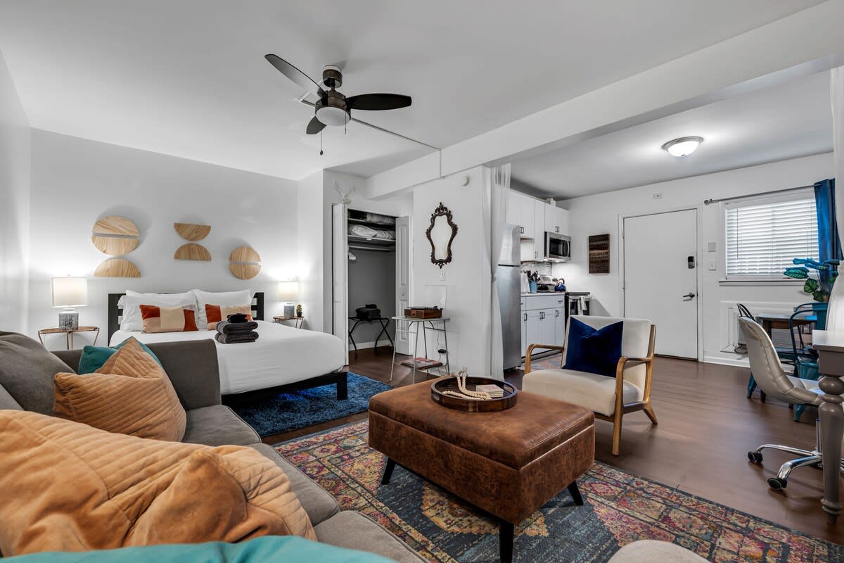 An open studio layout is presented, featuring a cozy seating area with a plush couch and a coffee table. A spacious bed is visible, paired with decorative wall elements. The kitchen area is equipped with modern appliances, and soft lighting enhances the overall ambiance.