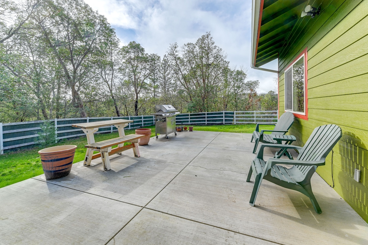 An outdoor patio area features a picnic table and two adirondack chairs, surrounded by greenery. A gas grill is positioned nearby, with a backdrop of a fenced yard and trees, creating a serene outdoor living space.