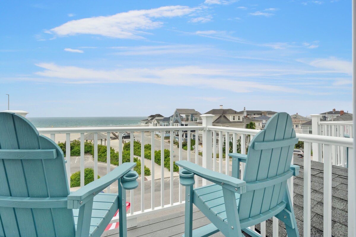 A private balcony is featured with two light blue adirondack chairs overlooking the ocean and sandy beach. The distant shore is lined with beach houses, while the clear blue sky creates a refreshing atmosphere.