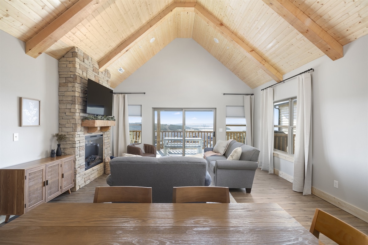 A spacious living area features vaulted wooden ceilings, enhancing the airy feel. A comfortable gray sofa and armchair are positioned in front of a stone fireplace, with large glass doors offering expansive views of the lake. Wooden dining furniture is visible in the foreground.