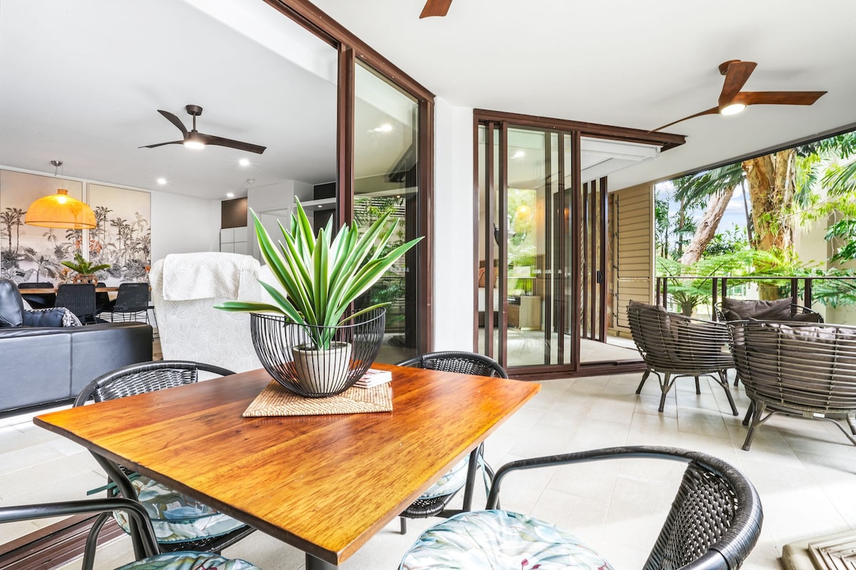 An inviting outdoor dining space is highlighted by a wooden table surrounded by four dark wicker chairs. Natural greenery and palm trees frame the view through expansive glass doors, while a cozy sitting area is visible in the background, featuring a neutral-toned sofa.