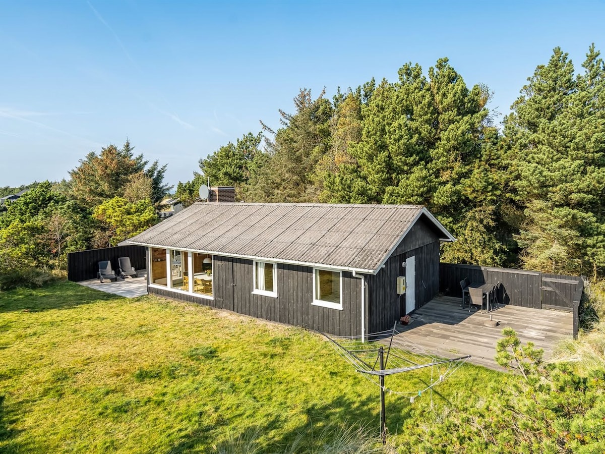 A single-story house is set amidst greenery, surrounded by trees. Large windows provide views of the outdoors. The building features a sloped roof, and a deck area is visible, furnished with seating for relaxation.
