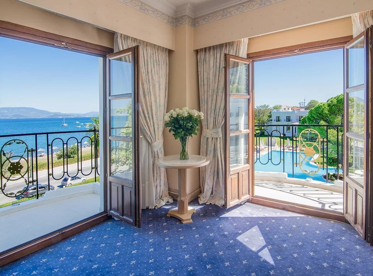 Large windows open to a scenic view of the sea and pool area. A round table with a vase of flowers is positioned centrally, while flowing curtains frame the view. The blue carpet features a subtle star pattern, enhancing the room's tranquil feel.