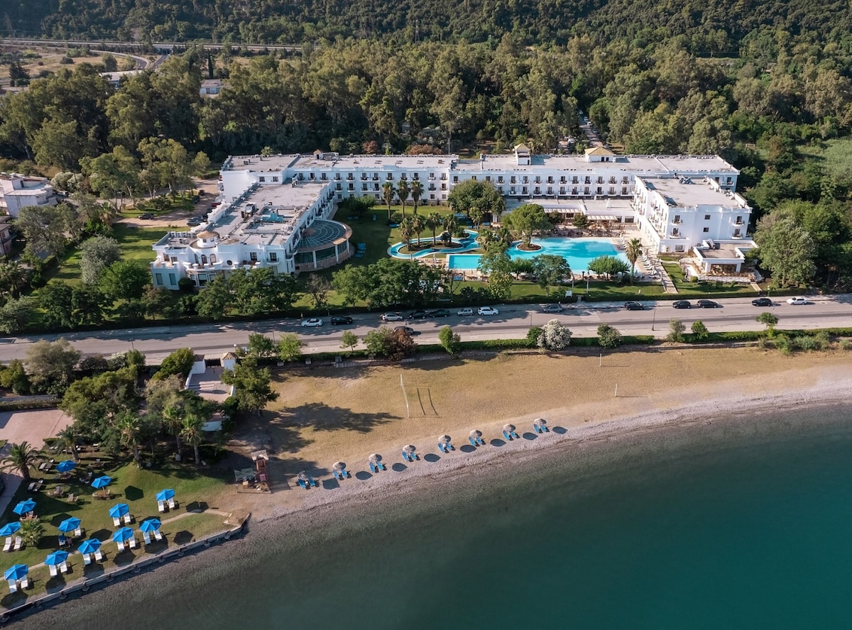 An aerial view captures a sprawling resort surrounded by lush greenery, featuring a large swimming pool and well-maintained gardens. Sun loungers with umbrellas line the sandy beach, enhancing the coastal setting. The building's architecture complements the serene landscape and highlights its inviting outdoor spaces.