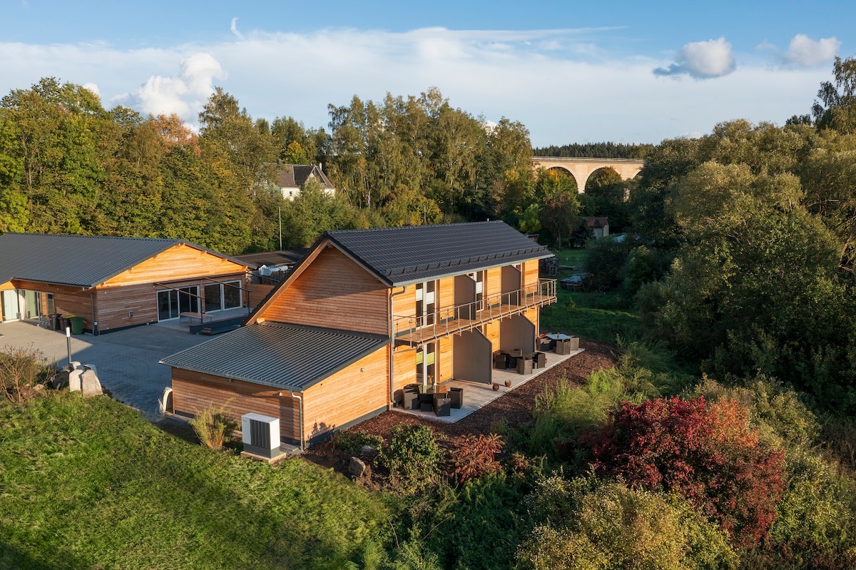 The two-story wooden structure is surrounded by greenery, featuring a covered balcony on the upper floor. Ample outdoor space is visible, with seating areas nestled within the landscaped surroundings. A bridge can be seen in the background, enhancing the natural setting.