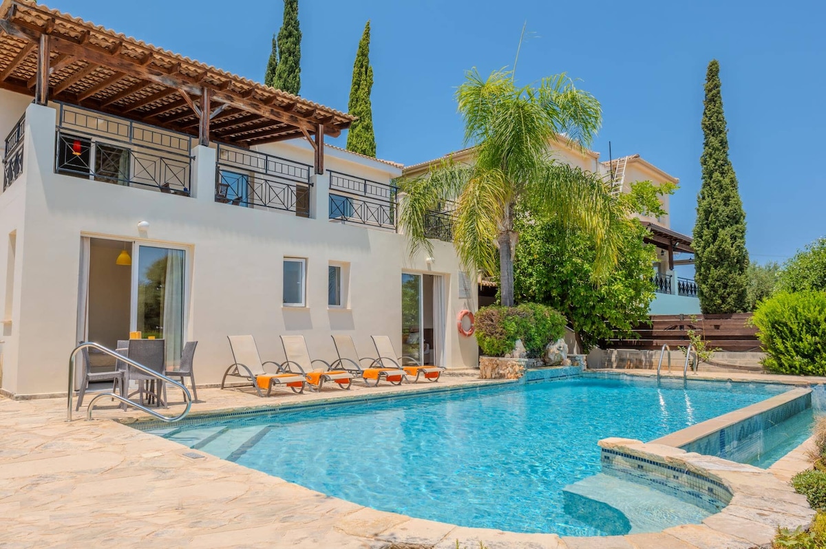 A large pool, measuring 10 x 5 meters, is situated in front of the villa, complete with a shallow entry area. Sun loungers are positioned around the pool, and the villa features multiple outdoor seating areas under a slatted pergola, providing shade and relaxation options.