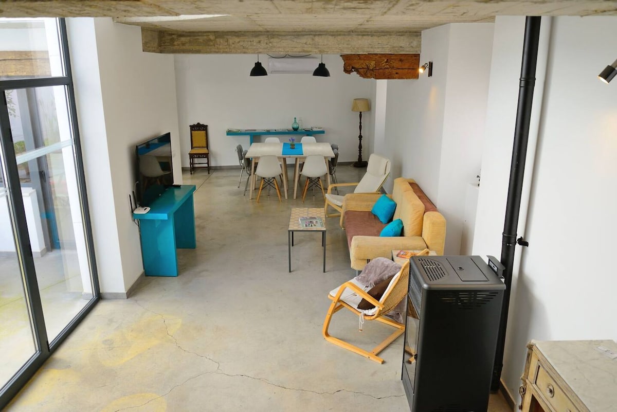 An open living space features a combination of modern and cozy furnishings. A wooden dining table is surrounded by six chairs, while a sofa and armchair provide comfortable seating. Large glass doors allow natural light and views of the outdoors, creating a bright atmosphere.