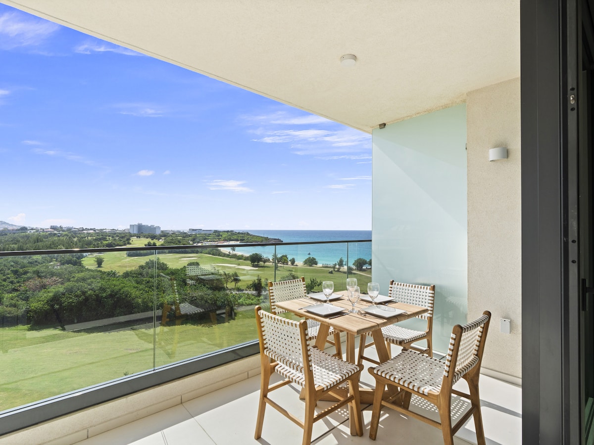 A private balcony features a wooden dining table surrounded by four woven chairs, offering a scenic view of the coastline and lush green landscape. The glass balustrade provides unobstructed visibility, allowing natural light to illuminate the space.
