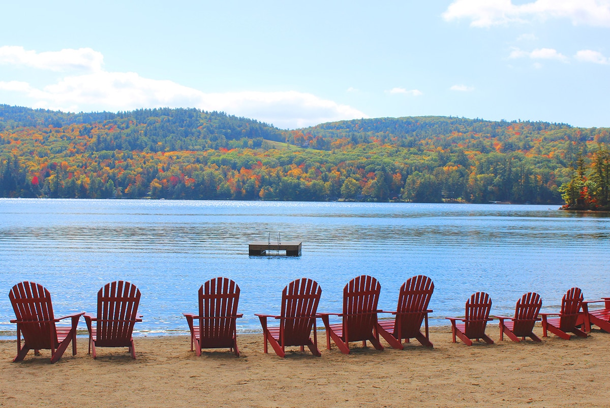 A sandy beach area is lined with a series of red Adirondack chairs facing a calm lake. The background is adorned with colorful autumn foliage on the hills, and a small dock is visible on the water.