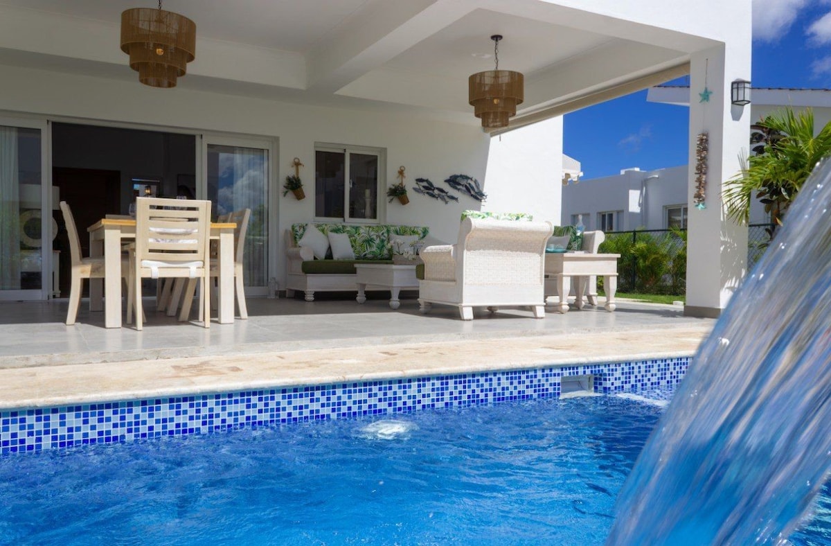 A private outdoor area features a sparkling pool with a cascading water feature. Comfortable seating and a dining table are provided under a covered terrace, creating a relaxing atmosphere. Sunlight filters through the space, emphasizing the serenity of the surroundings.