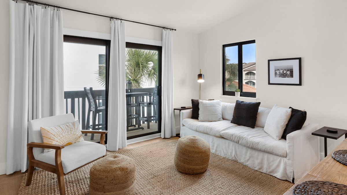 A bright living area features a cozy white sofa with dark accent pillows, complemented by a pair of round ottomans. Natural light floods the space through large windows, revealing the outdoor balcony. Sheer white curtains frame the windows, adding a touch of elegance.