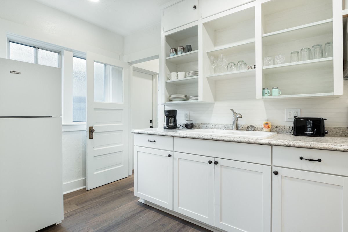 The kitchen features white cabinetry and a granite countertop, complemented by full-size appliances including a refrigerator and coffee maker. Open shelving displays glassware, while the space is illuminated by natural light coming through a nearby window and door.