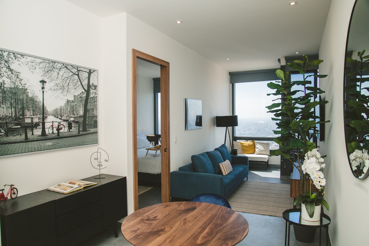 A well-lit living area features a blue sofa positioned near a large window, offering views of the city. A modern round dining table made of wood complements the space, and a tall plant adds a touch of greenery. Artistic wall decor enhances the ambiance.