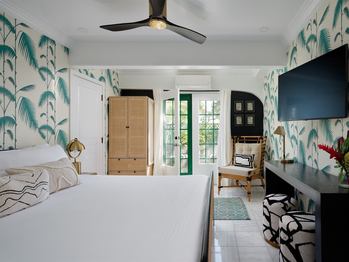 The spacious room features tropical-themed wallpaper and a king-sized bed. A seating area is included with a textured chair and a bamboo-style accent chair. French doors provide access to a private patio, with soft light entering through the draped curtains.