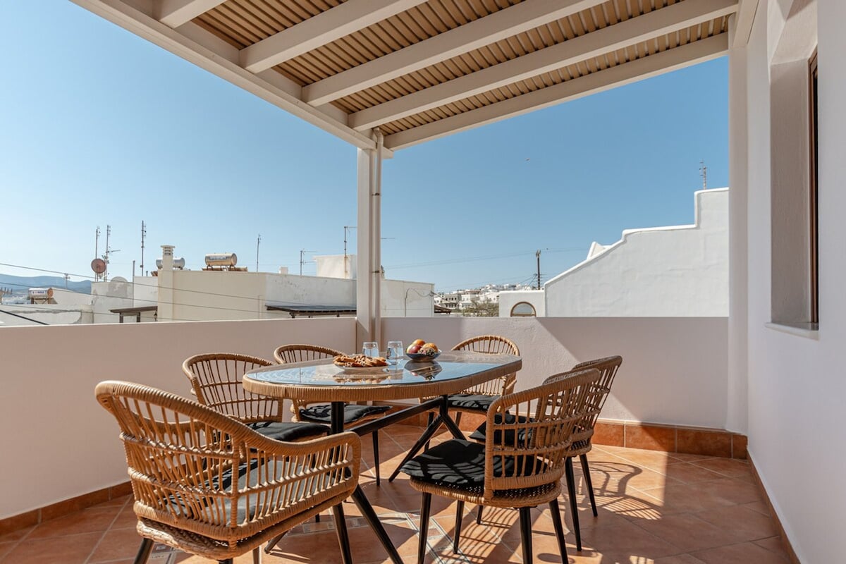 A spacious terrace is presented with a round table surrounded by four wicker chairs, offering a comfortable dining space. The area features a tiled floor and is sheltered by a wooden overhang. Clear views of the white-washed buildings in the distance are visible under a bright blue sky.