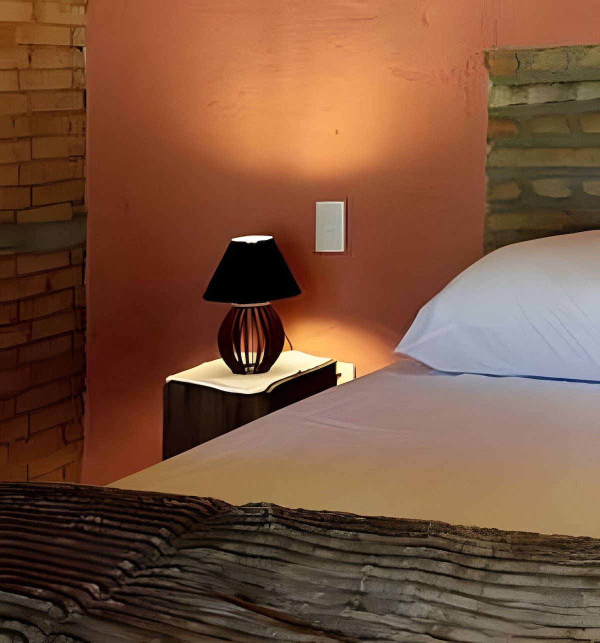 A cozy corner of the bedroom is highlighted by a nightstand featuring a stylish lamp. The spacious bed is adorned with soft linens, while the warm hues of the wall introduce a sense of tranquility to the rustic space.