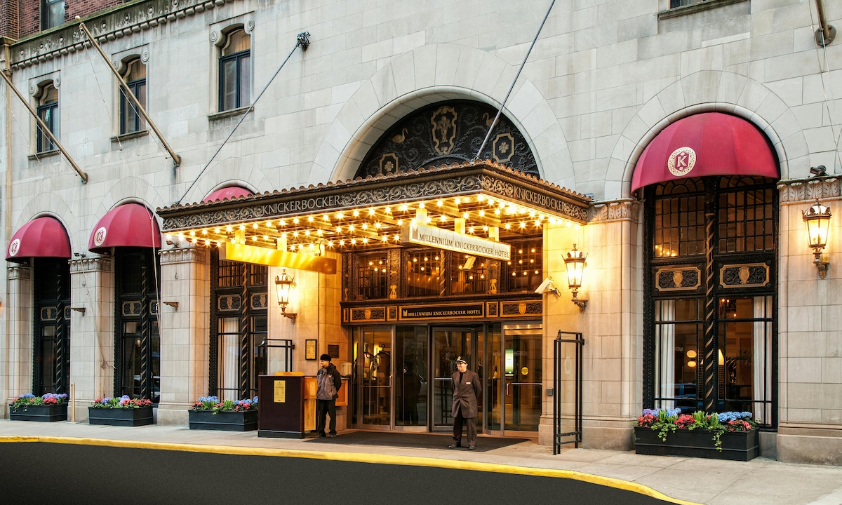 The hotel's entrance is adorned with vintage-style lighting and grand awnings. Flanking the front door are broad windows, enhancing the inviting appearance. Decorative flower boxes add a touch of elegance, while the building's architectural details reflect its historical significance.