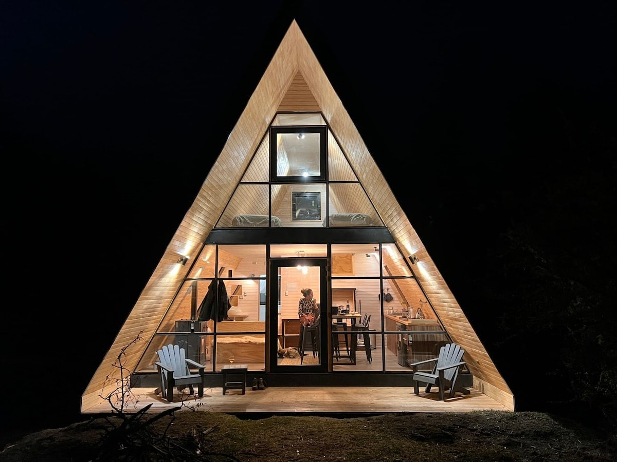 The triangular cabin structure is illuminated at night, revealing large windows that provide a view into the welcoming interior. Adirondack chairs are positioned on the porch, inviting guests to enjoy the peaceful surroundings. Soft light enhances the warm atmosphere of the open space inside.