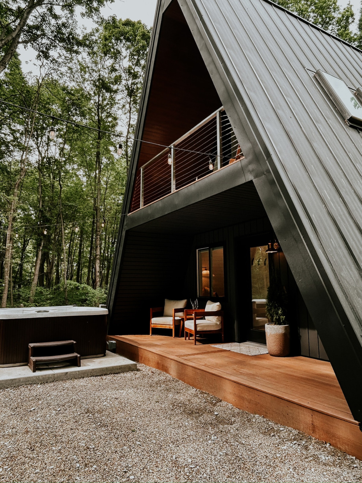 The modern A-frame cabin features a striking sloped roof, complemented by a wooden deck. Comfortable seating is arranged on the deck, inviting relaxation. A hot tub is positioned nearby, surrounded by natural greenery, enhancing the tranquil outdoor space.