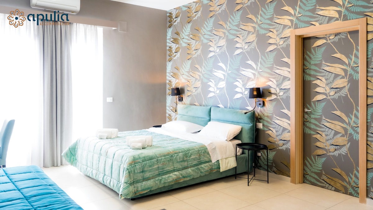 A modern sleeping area features a spacious bed with soft bedding and large, decorative pillows. The walls are adorned with a botanical pattern, complemented by stylish sconces. Natural light streams in through a floor-to-ceiling window, enhancing the serene ambiance.
