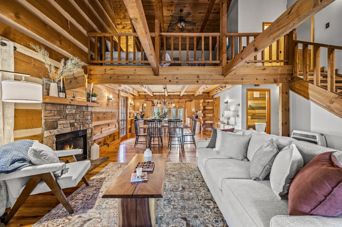 An open living area showcases vaulted tongue and groove ceilings supported by wooden beams. A cozy gas fireplace is framed by a natural stone surround, while a comfortable couch and armchair offer seating. The space is illuminated by soft lighting and features a large dining table in the background.