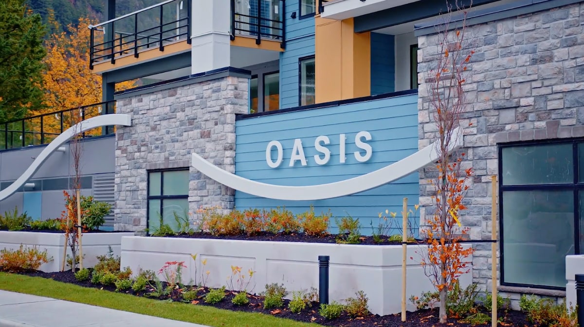 The exterior of the building displays a modern design, featuring large windows and a combination of stone and blue siding. The sign 'OASIS' is prominently featured above the entrance. Seasonal greenery and colorful foliage are present in the landscaped area below.