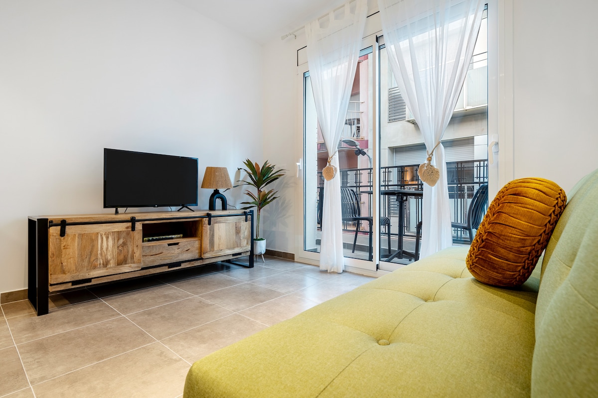 A sunlit living area features a green sofa bed and a wooden television stand. A TV and a decorative lamp are part of the furnishings. Floor-to-ceiling glass doors lead to a small balcony, letting in natural light and offering a view of the outside.