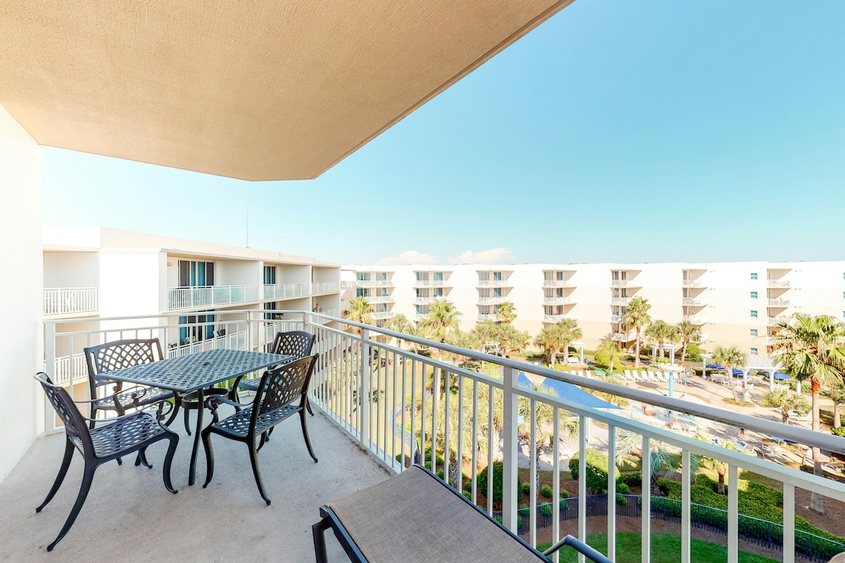 A private balcony view is showcased, featuring a table and chairs positioned for relaxation. Below, a landscaped area with palm trees and a glimpse of the pool area can be seen, creating a sense of connectivity to the outdoor surroundings.
