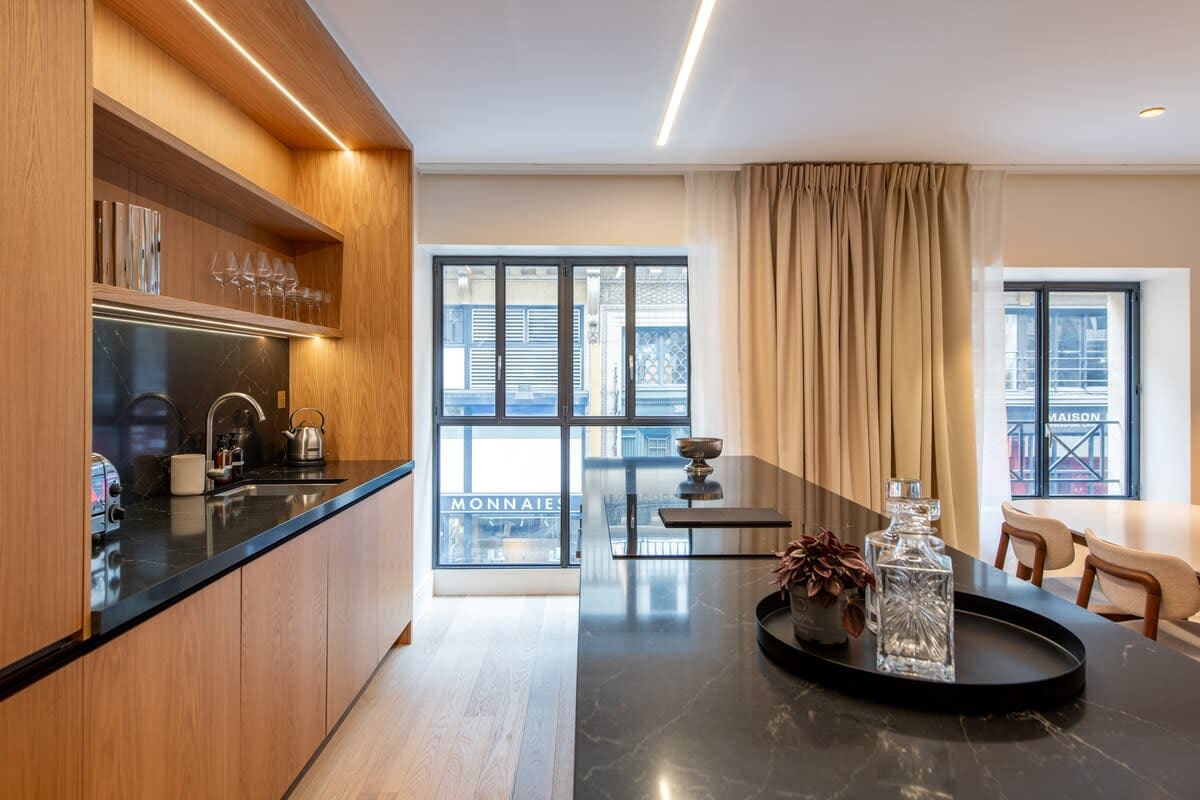 A modern kitchen area features wooden cabinetry with glassware displayed above. A large marble kitchen island provides ample space for dining, with elegant decor items placed atop it. Natural light filters through large windows adorned with soft curtains, enhancing the bright and airy ambiance.