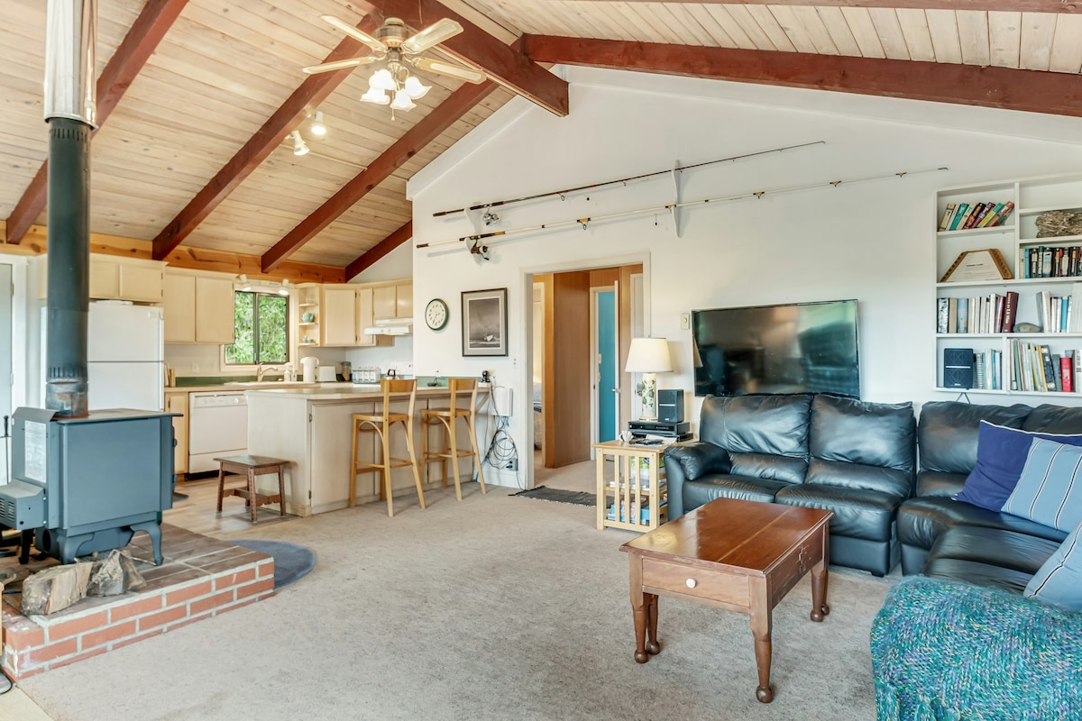 An open living area features vaulted wooden ceilings and a wood stove. A dark leather sofa is positioned around a wooden coffee table. A flat-screen television is mounted on the wall, while a kitchen area with bar seating is visible in the background.