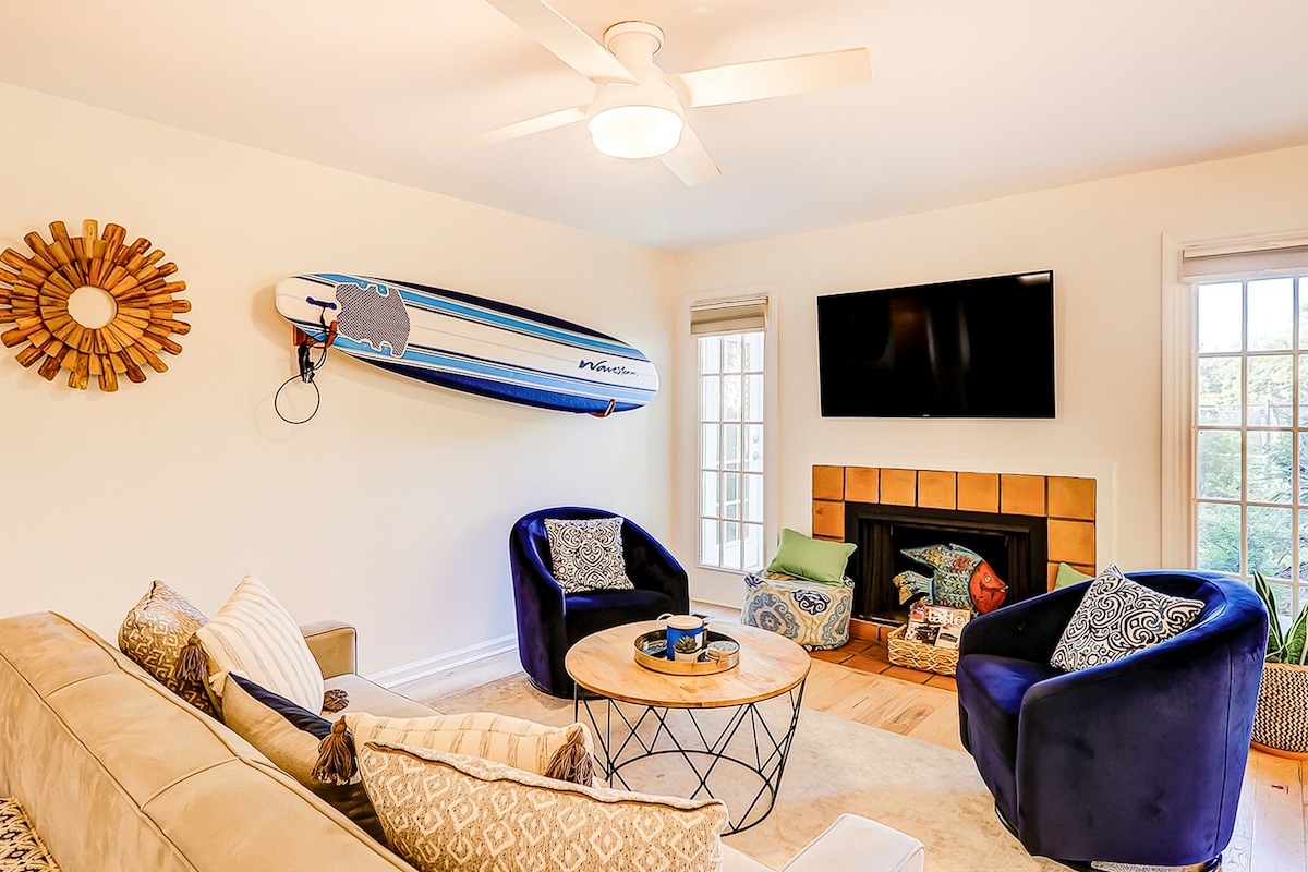 The main living area features a comfortable sectional sofa and a pair of blue accent chairs surrounding a circular coffee table. A TV is mounted above a decorative fireplace, and a surfboard is mounted on the wall, adding a relaxed coastal touch. Natural light enters through the windows.
