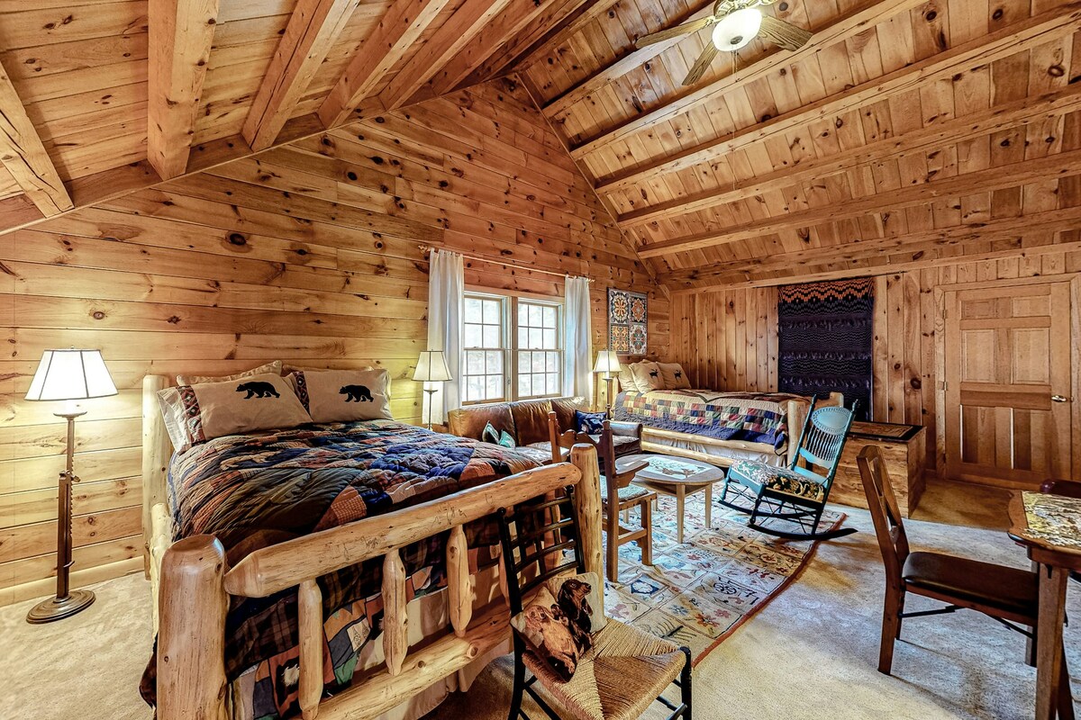 A rustic bedroom setting features log cabin walls adorned with natural wood. Two beds are dressed in quilts, accompanied by a rocking chair and a small table. Light fixtures provide gentle illumination, while windows allow for natural light and views of the trees outside.