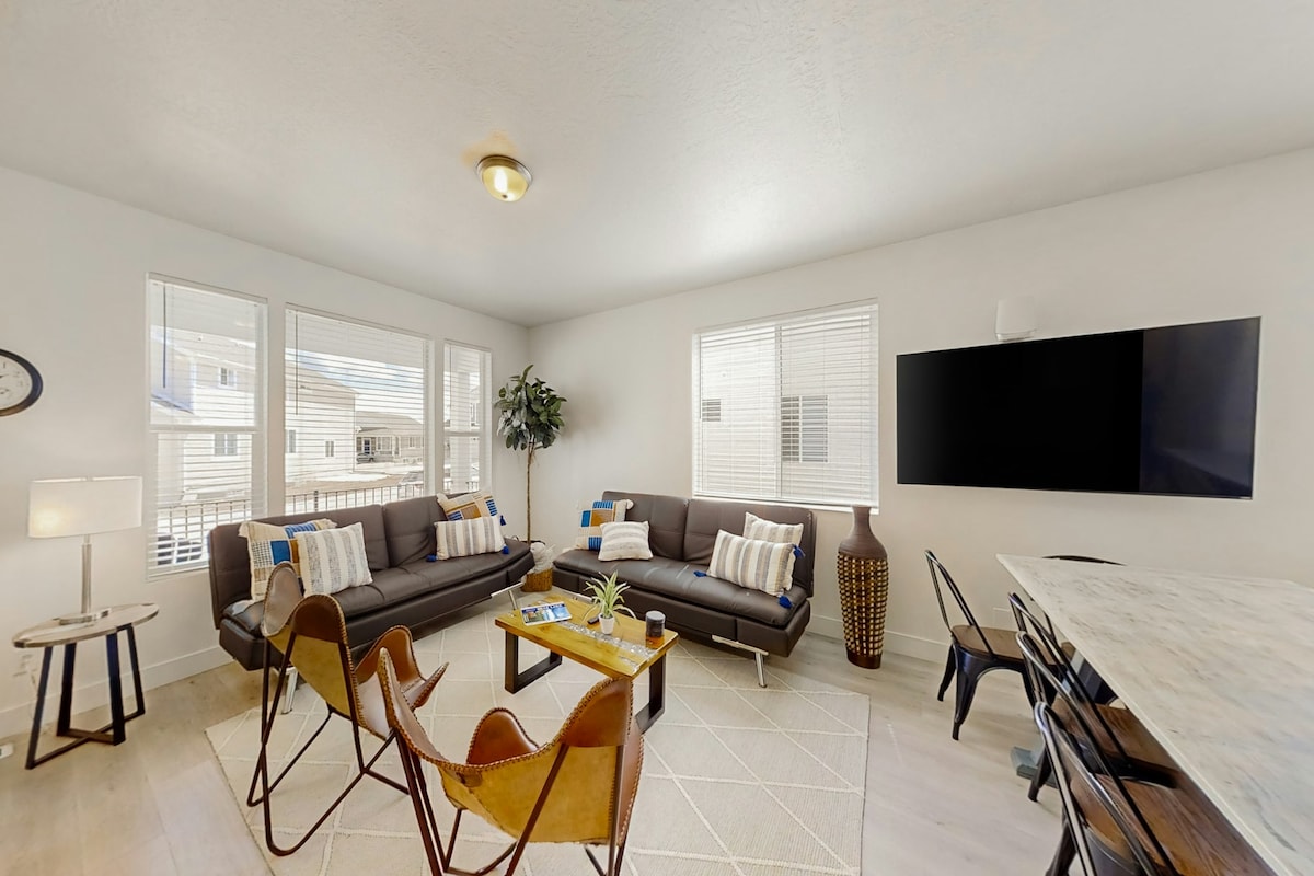 The open-concept living area features comfortable seating arrangements with dark sofas and modern accent chairs. Natural light filters through the multiple windows, illuminating the space. A flat-screen TV is mounted on the wall opposite a marble dining table, creating a welcoming environment for relaxation and socializing.