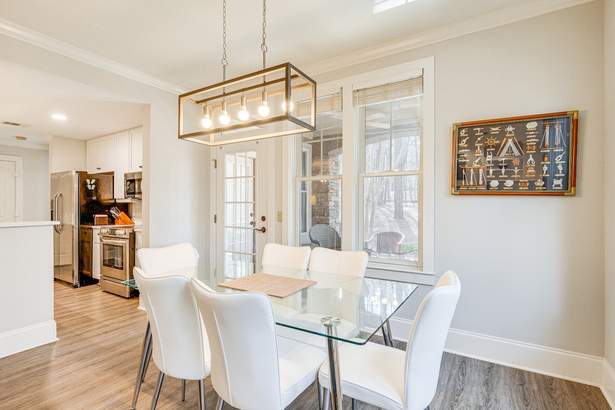 A dining area features a modern glass table surrounded by six sleek white chairs. Natural light fills the space through large windows, and a stylish light fixture hangs above the table. In the background, sliding glass doors lead to an outdoor patio.