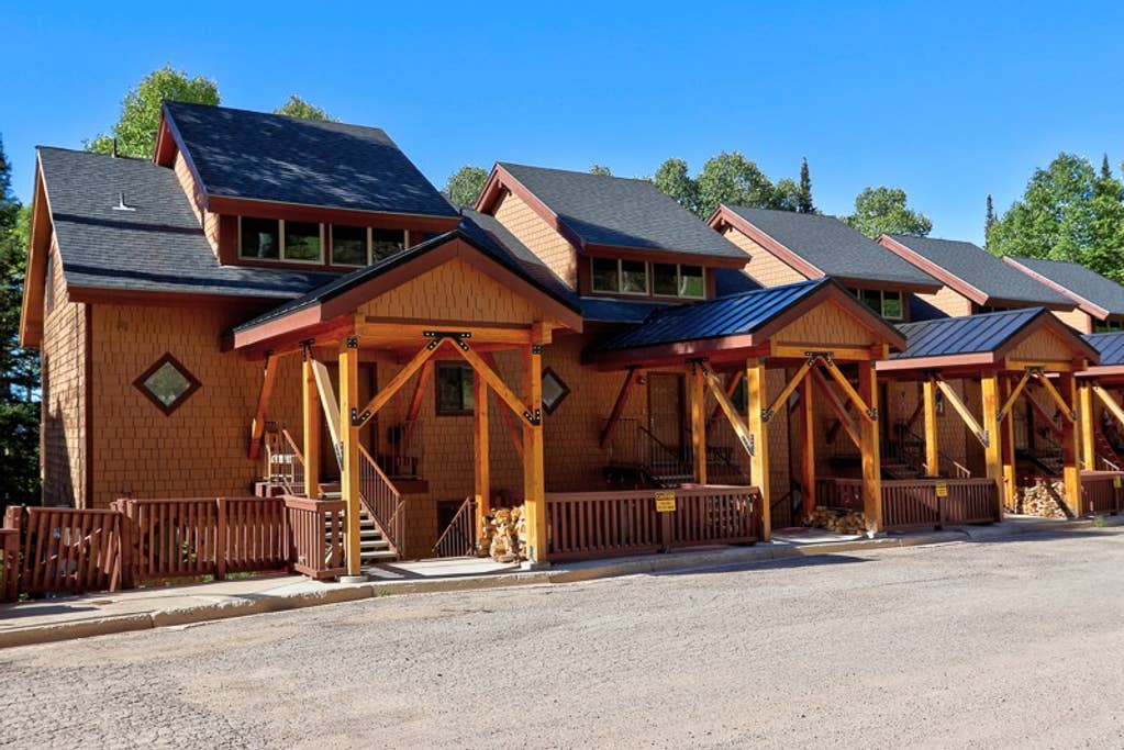 A row of charming mountain cabins features wood-paneled exteriors and gabled roofs. Each cabin is supported by sturdy columns and includes a set of steps leading to the entrance. Surrounding greenery adds to the serene setting of the Eagle Point Ski Resort.