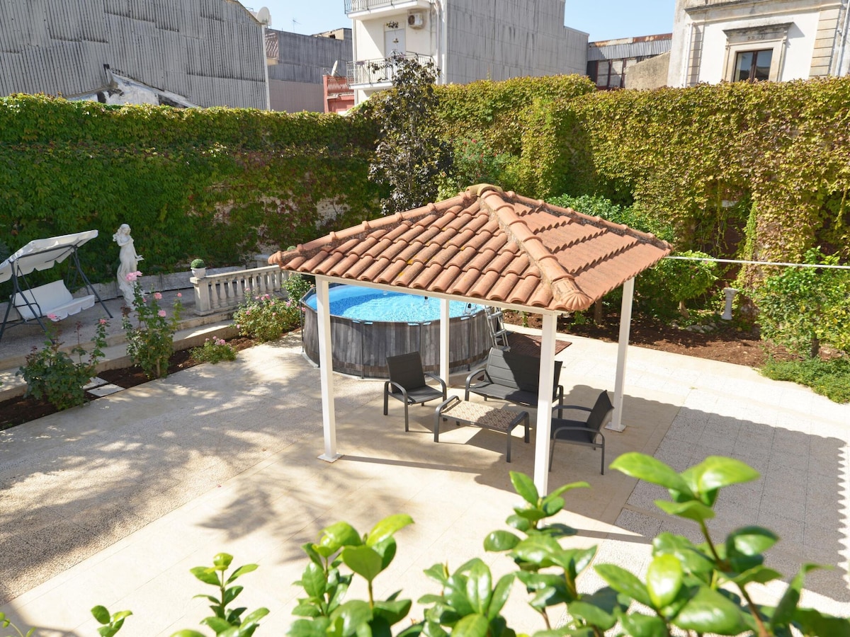 An outdoor pavilion features a tiled roof and is furnished with seating for relaxation. The small above-ground pool is visible nearby, surrounded by well-maintained greenery, including shrubs and trees, creating a serene outdoor space.