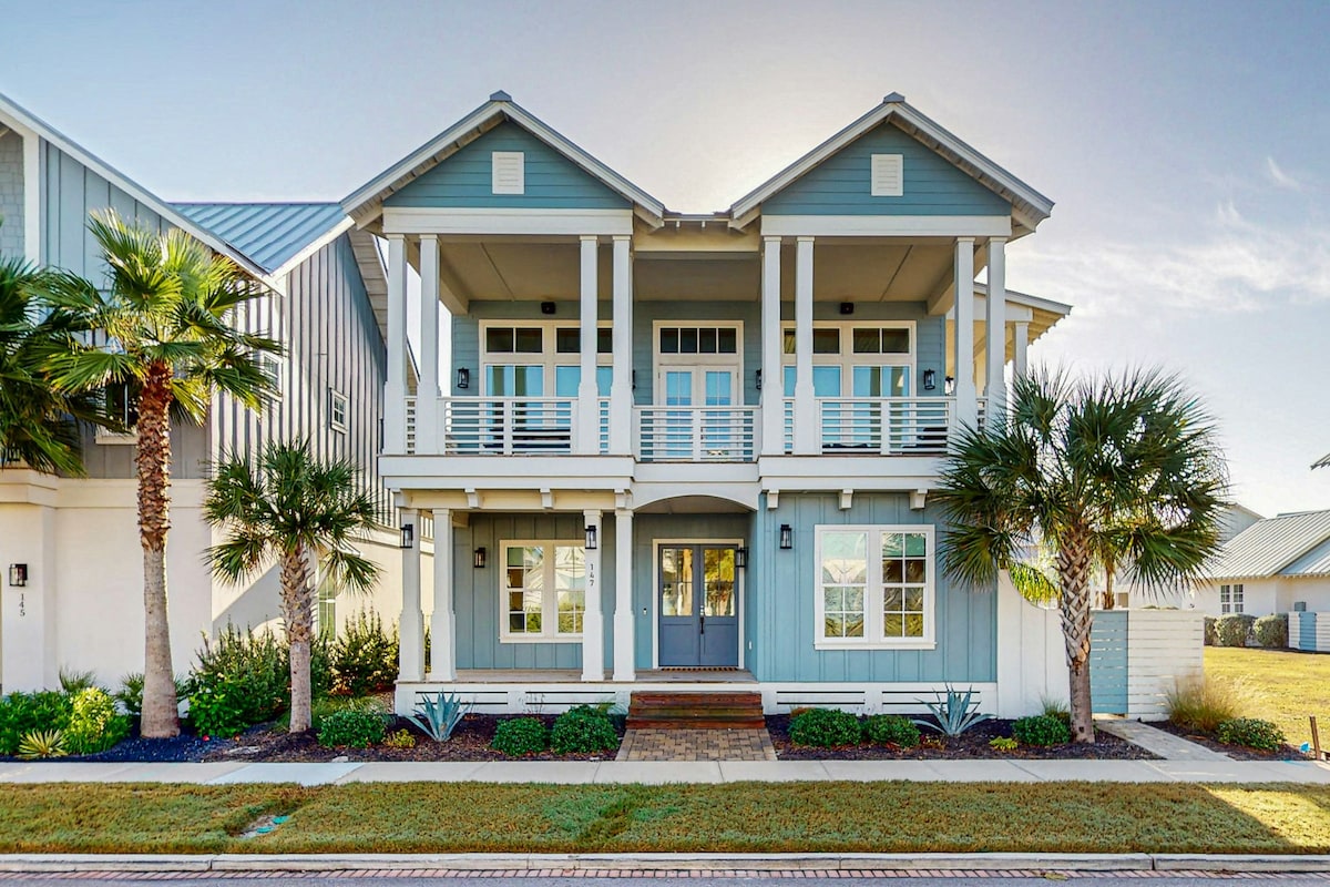 The exterior of the home features a two-story design with a light blue facade and welcoming front porch. Palm trees and lush greenery surround the entrance, which includes a stone pathway leading to a central door framed by large windows.