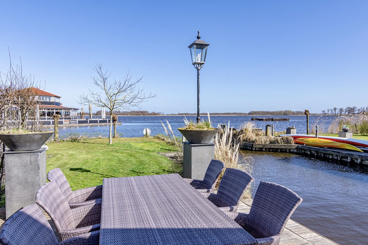 An outdoor dining area is presented with a large table surrounded by six wicker chairs. The setting overlooks a serene waterway, complemented by a vintage lamp post and lush green grass. A colorful boat is visible along the edge of the water.