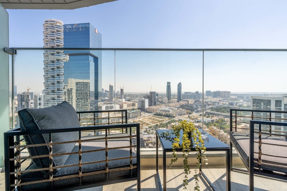 A balcony offers seating with modern chairs, overlooking a panoramic view of Dubai's skyline. The space is complemented by a small table adorned with greenery, providing a serene spot to enjoy the surroundings with clear skies in the background.