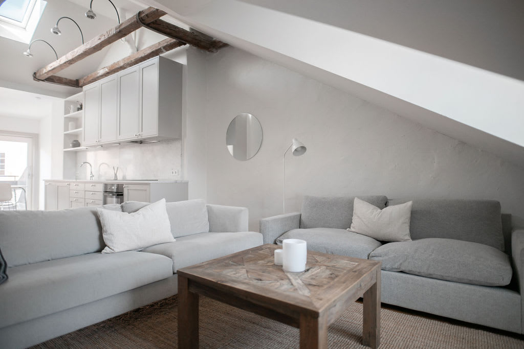A cozy living area is presented with two spacious gray sofas adorned with decorative pillows. A wooden coffee table occupies the center, while soft light is provided by a modern floor lamp. The kitchen area is visible in the background, featuring sleek cabinetry.