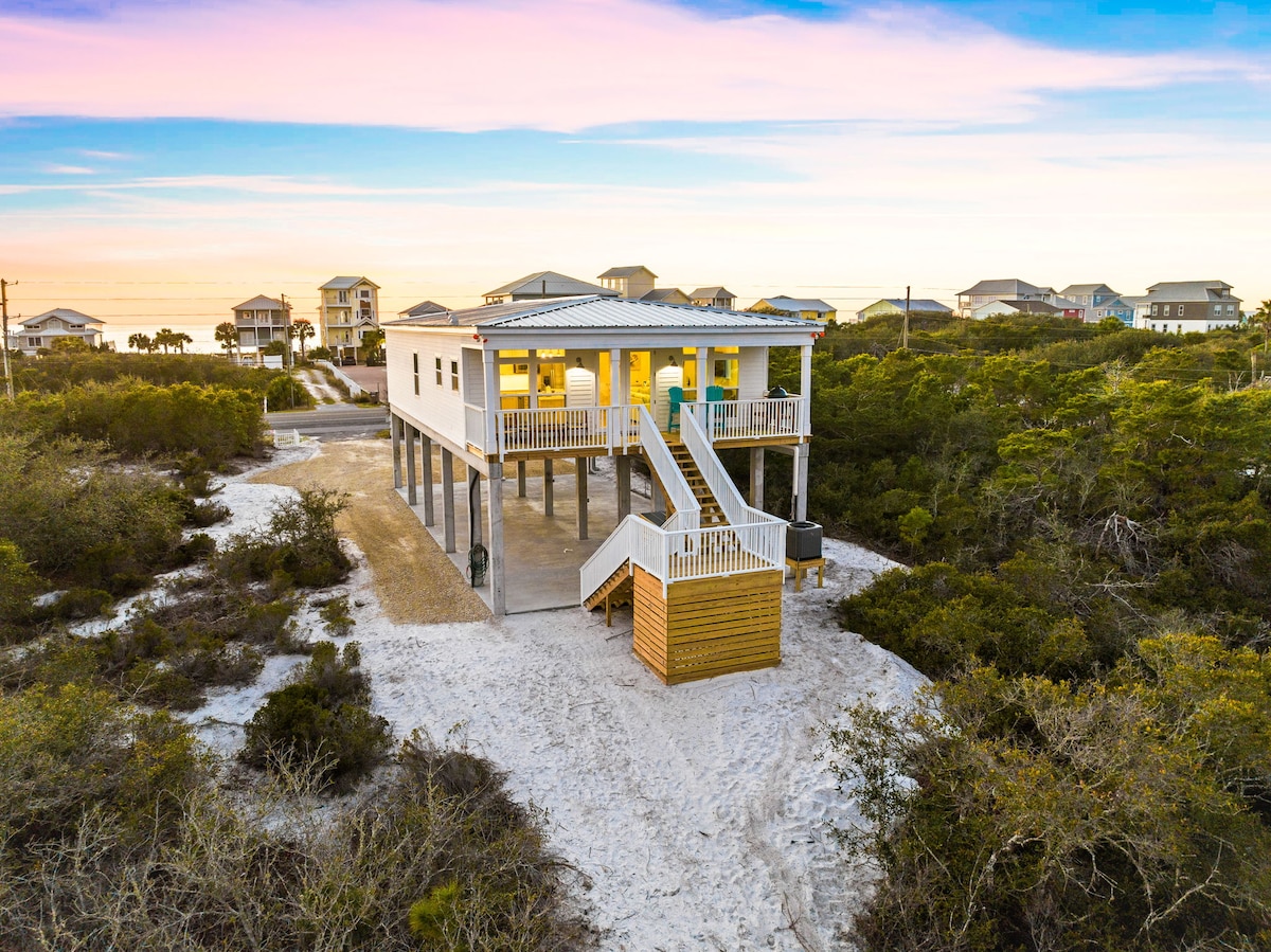 The elevated exterior of the home is visible, showcasing a spacious staircase leading to the front porch. Surrounding greenery and sandy pathways offer a natural setting. The home is positioned away from neighboring structures, contributing to a sense of privacy.