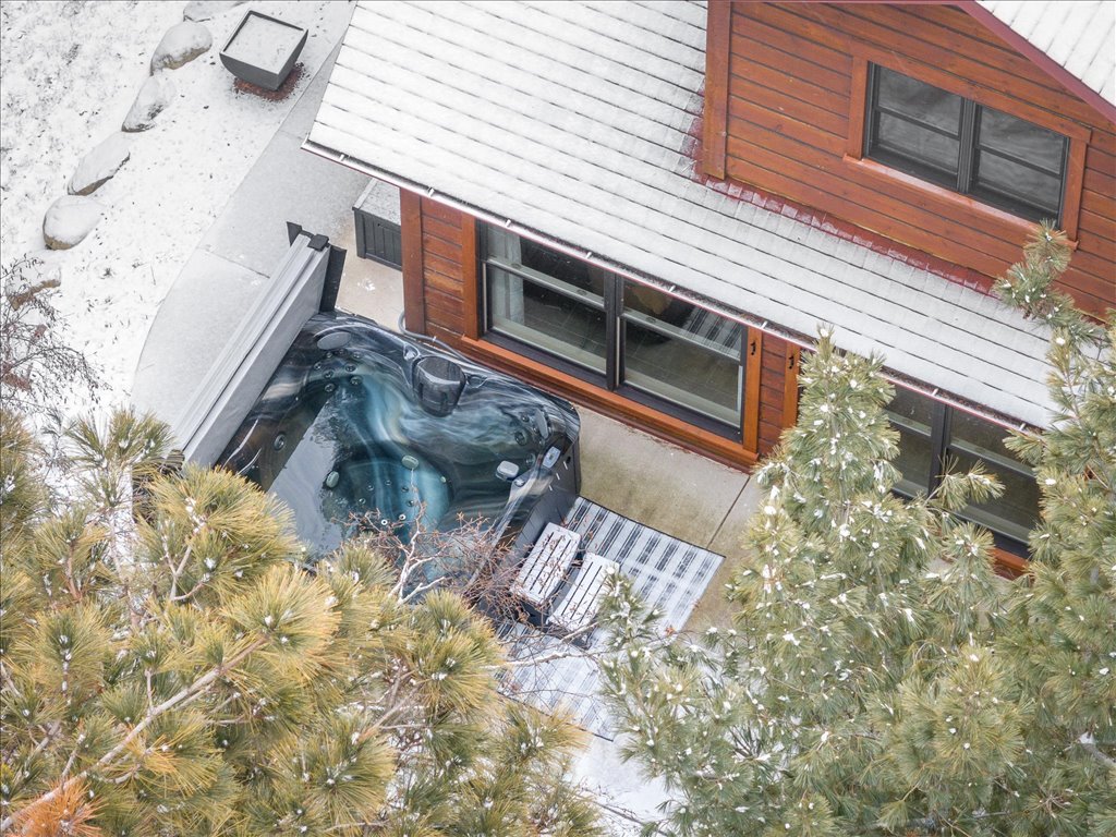 An aerial view of the cabin reveals a private hot tub situated on a concrete patio. Surrounding pine trees create a natural frame, while a soft layer of snow covers the ground, adding a seasonal touch. The cabin features large windows, allowing ample light into the space.