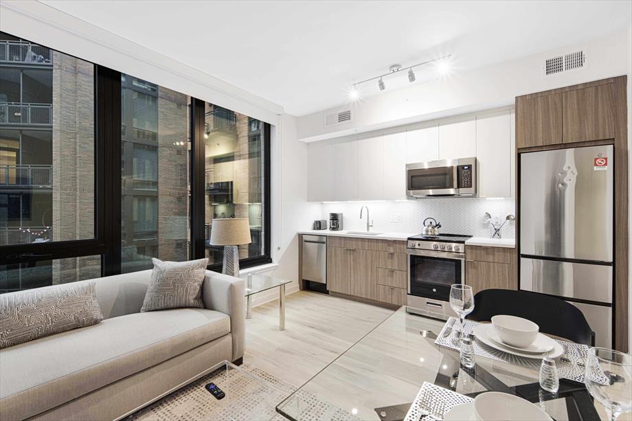 A modern living area is showcased with a comfortable light-colored couch positioned adjacent to a glass dining table set for four. The kitchen features sleek cabinetry, stainless steel appliances, and ample natural light from large windows overlooking the lively neighborhood.