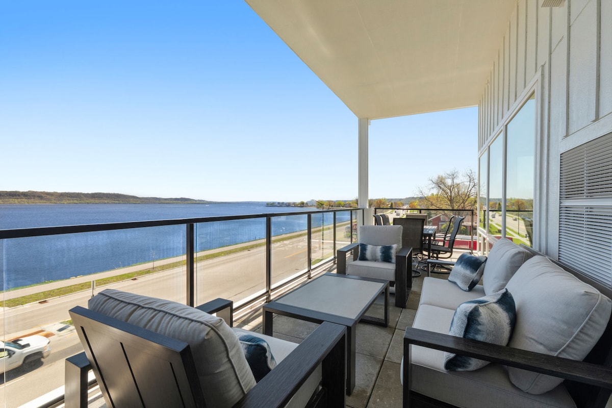 A private balcony features comfortable outdoor seating arrangements, including a sofa and chairs. The space overlooks tranquil waters, providing expansive views of the lake and surrounding landscape under a clear blue sky.