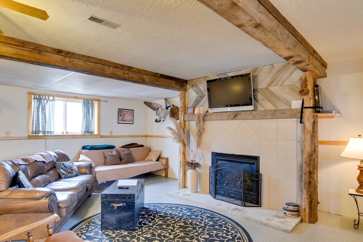 The living area features a leather sofa and a full futon, complemented by rustic wooden beams overhead. A flat-screen TV is mounted on a patterned wall, above an elegant gas fireplace set within a stone surround. A patterned area rug enhances the cozy atmosphere.