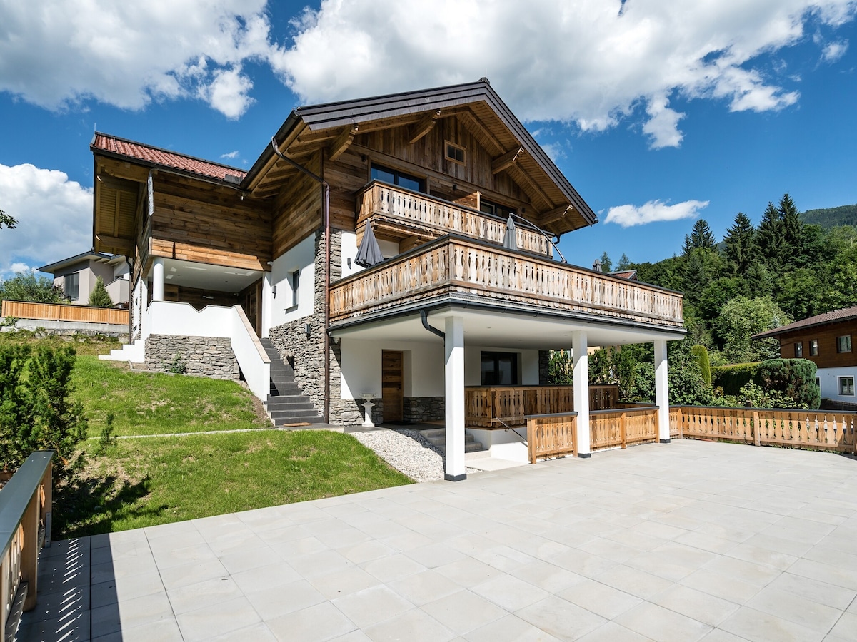 A spacious holiday home is presented from an angled view, showcasing its wooden balconies and distinct architectural elements. Well-maintained landscaping surrounds the property, and a covered patio area can be seen at the ground level. Blue skies and fluffy clouds create a welcoming atmosphere.